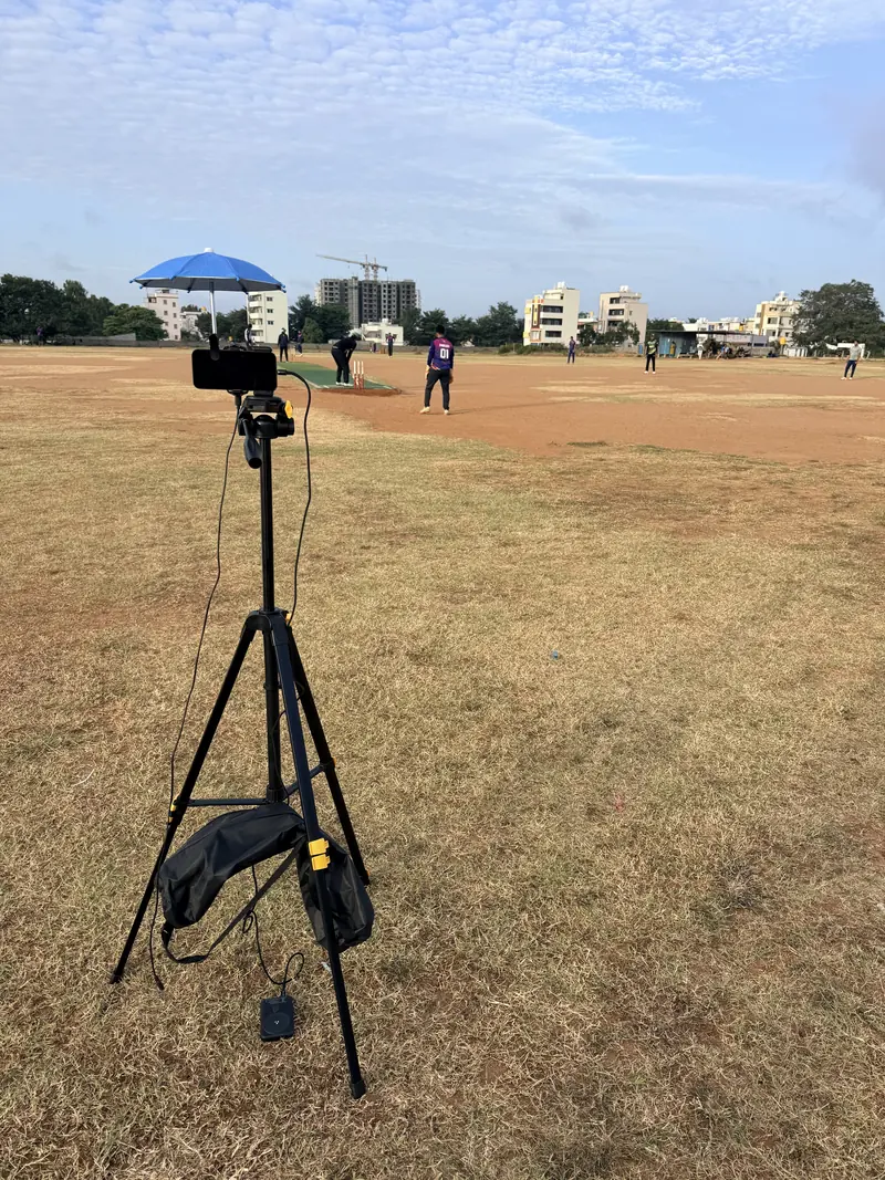 A smartphone on a tall tripod behind the wicketkeeper at a local cricket ground, shaded by a clip-on blue umbrella, with a power bank hanging from the tripod and a match in progress on the pitch ahead