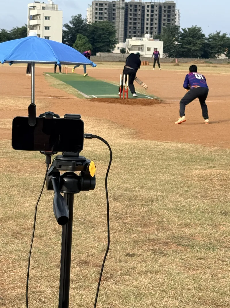 Close-up of the streaming phone mounted on a tripod pan head, fully shaded by a clip-on blue umbrella, with the charging cable running to a power bank and the pitch, batter and wicketkeeper visible ahead